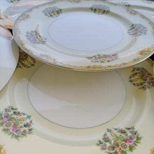 May include: A set of vintage porcelain plates. The plates feature a cream-coloured base, gold rims, and floral patterns in pink, blue, and green. The plates are stacked, highlighting their decorative elements.