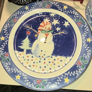 Op de afbeelding: Een blauwe en witte keramische dinerbord met een sneeuwpopontwerp. De sneeuwpop draagt ​​een rood-witte hoed en een groene sjaal. Het bord heeft een blauwe rand met een witte wijnstok en gele sterren.