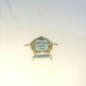 May include: A white porcelain plate with a decorative emblem in the centre. The emblem features the text "NORITAKE CHINA JAPAN" in a light blue oval, surrounded by gold and floral designs, with the word "CLAREMONT" below.