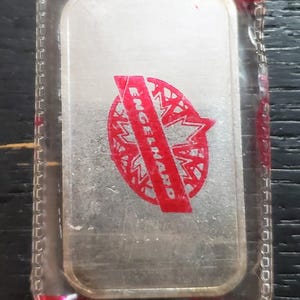 Puede incluir: Barra de plata dentro de un estuche de plástico transparente. La barra tiene un logotipo rojo que dice "Engelhard". El estuche está sellado con cinta roja.