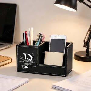 May include: Black desk tidy with white stitching, holding pens, a smartphone, and a notepad. The organiser features the letter "D" with the name "Sophia" below it, in white lettering. The organiser is on a desk with a laptop and lamp.