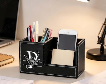 May include: Black desk organizer with white stitching, holding pens, a smartphone, and a notepad. The organizer features the letter "D" with the name "Sophia" below it, in white lettering. The organizer is on a desk with a laptop and lamp.