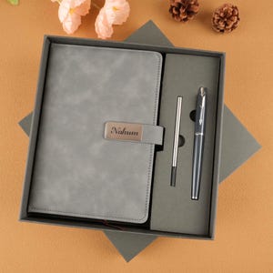 Könnte beinhalten: Ein graues, ledergebundenes Notizbuch mit einem Metallverschluss mit dem Namen "Nahum" wird in einer Geschenkbox präsentiert. Ein passender Stift und eine Mine sind enthalten. Das Set wird auf einer braunen Oberfläche mit dekorativen Blumen und Tannenzapfen präsentiert.