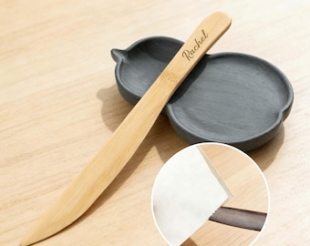 May include: A light-colored bamboo letter opener with the name "Rachel" engraved on the handle. The opener rests on a dark gray, leaf-shaped dish. A close-up shows the edge of a white paper.