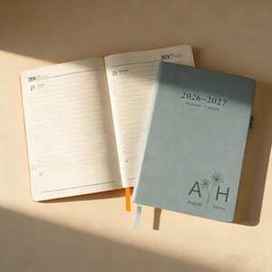 Op de afbeelding: Een open oranje notitieboek naast een gesloten lichtblauwe 2026-2027 maandplanner. De planner heeft de tekst "2026-2027 Monthly Planner" en de initialen "AH" met de namen "August" en "Henry" eronder.