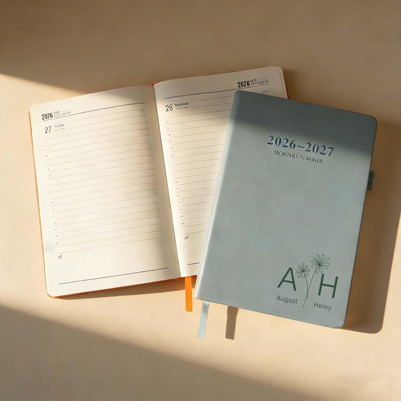 Op de afbeelding: Een open oranje notitieboek naast een gesloten lichtblauwe 2026-2027 maandplanner. De planner heeft de tekst "2026-2027 Monthly Planner" en de initialen "AH" met de namen "August" en "Henry" eronder.