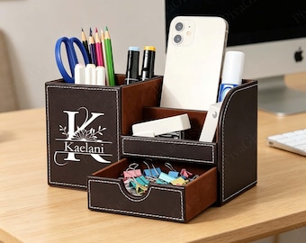 Op de afbeelding: Een bruine leren bureau-organizer met meerdere compartimenten. Het bevat een schaar, potloden, stiften, een telefoon en andere kantoorbenodigdheden. De organizer heeft een lade met paperclips en de naam "Kaelani" staat erop.