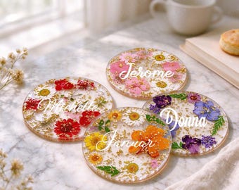 May include: Four circular resin coasters featuring embedded pressed flowers in red, pink, white, yellow, orange, and purple, accented with gold flakes. Each coaster displays a white cursive name: Michelle, Jerome, Januar, and Donna.