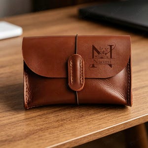 May include: A brown leather wallet featuring a flap closure secured by a vertical leather strap and an elastic cord. The front is engraved with a large letter N, a floral design, and the name Naomi in a dark, clear, and elegant font style.