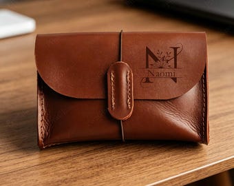 May include: A brown leather wallet featuring a flap closure secured by a vertical leather strap and an elastic cord. The front is engraved with a large letter N, a floral design, and the name Naomi in a dark, clear, and elegant font style.