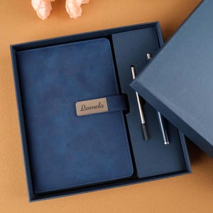 May include: A blue leather-bound journal with a silver nameplate that reads "Pamela" is in a blue gift box with a silver pen.