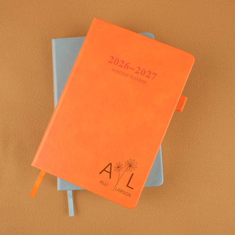 Op de afbeelding: Een oranje maandplanner voor 2026-2027 met een bijpassende oranje lint-bladwijzer. De omslag bevat de tekst "2026-2027 MONTHLY PLANNER" en de initialen "A L" met bloemaccenten, en de naam "Alli Larson". Een lichtblauwe planner is eronder zichtbaar.