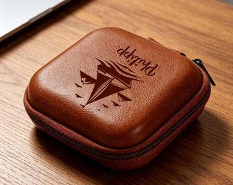 May include: A square, brown, textured faux leather zippered case featuring a dark brown engraved graphic of a sailboat on the water with mountains in the background and the name Philipp written in a cursive script above the sailboat design.