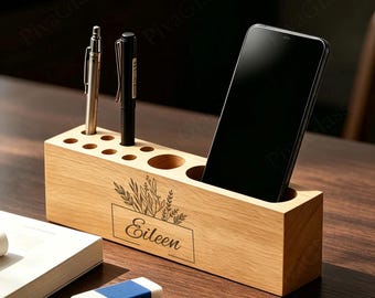 May include: A rectangular light-colored wooden desk organizer featuring multiple circular holes for pens and a wider slot holding a black smartphone. The front is engraved with a botanical illustration and the name Eileen in cursive script.