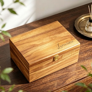 May include: A rectangular wooden box with a natural grain finish, featuring a gold-coloured metal latch on the front. The lid is engraved with the name Jamarino in a cursive script. The box has a hinged lid and a smooth, polished surface.