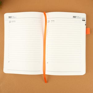 Op de afbeelding: Open planner met gelinieerde pagina's, gedateerd 2027. De linkerpagina toont woensdag 20 januari en de rechterpagina toont donderdag 21 januari. Een oranje lint-bladwijzer staat in het midden. De planner ligt open op een bruin oppervlak.