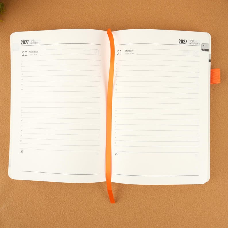 Op de afbeelding: Open planner met gelinieerde pagina's, gedateerd 2027. De linkerpagina toont woensdag 20 januari en de rechterpagina toont donderdag 21 januari. Een oranje lint-bladwijzer staat in het midden. De planner ligt open op een bruin oppervlak.