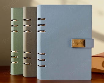 May include: Three leather-bound journals in shades of green, gray, and blue. Each journal features a metal clasp closure and a series of metal rings along the spine. The blue journal has a gold nameplate that reads "Angelika".