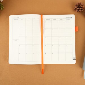 Op de afbeelding: Open witte planner met een maandelijkse kalenderindeling, met een oranje lint-bladwijzer. De planner is open op een decemberpagina, met de datum 12 zichtbaar. De achtergrond is effen bruin.
