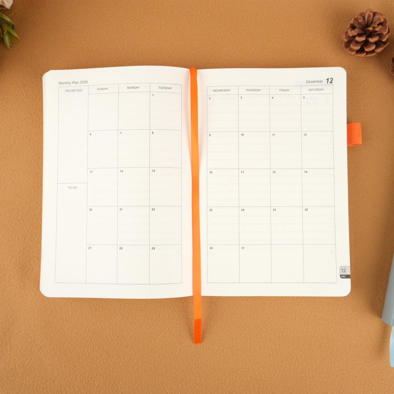 Op de afbeelding: Open witte planner met een maandelijkse kalenderindeling, met een oranje lint-bladwijzer. De planner is open op een decemberpagina, met de datum 12 zichtbaar. De achtergrond is effen bruin.