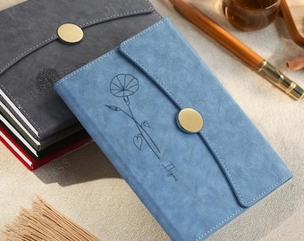 May include: Two faux-suede notebooks, one gray and one blue, with gold button closures. The blue notebook features a floral design and the word "Ilyas". The gray notebook has a tree design. Both have stitched edges.