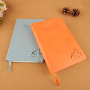 Op de afbeelding: Twee hardcover maandplanners, &eacute;&eacute;n lichtblauw en &eacute;&eacute;n oranje. De oranje planner heeft de tekst "2026-2027 Monthly Planner" en de initialen "AL" met de naam "Alli Larson". De lichtblauwe planner heeft de tekst "2026 Monthly Planner" en de initialen "AM" met de naam "Emma Melville".