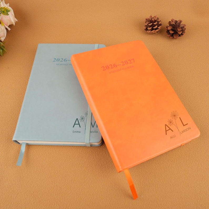 Op de afbeelding: Twee hardcover maandplanners, &eacute;&eacute;n lichtblauw en &eacute;&eacute;n oranje. De oranje planner heeft de tekst "2026-2027 Monthly Planner" en de initialen "AL" met de naam "Alli Larson". De lichtblauwe planner heeft de tekst "2026 Monthly Planner" en de initialen "AM" met de naam "Emma Melville".