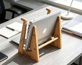 May include: A light-colored bamboo laptop stand with a vertical support structure and a horizontal crossbar. The stand features the name Shaun engraved on the side panel and holds a silver laptop with a visible Apple logo on the back casing.
