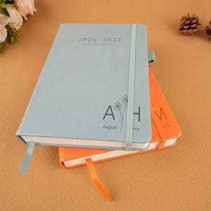 Op de afbeelding: Twee notitieboeken, &eacute;&eacute;n lichtblauw en &eacute;&eacute;n oranje, zijn op elkaar gestapeld. Op het lichtblauwe notitieboek staat de tekst "2026-2027 MONTHLY PLANNER" en "August". Beide notitieboeken hebben een lint als bladwijzer. Op een bruin oppervlak.
