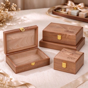 May include: Three rectangular wooden boxes with natural wood grain patterns. Two boxes are closed with gold-coloured metal latches and feature engraved names, Michelle and WENDY. One box is open, revealing a light-coloured interior wood finish.