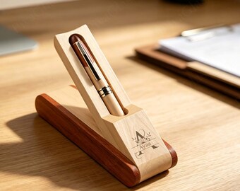 May include: A light-colored wooden pen case with a slanted top slot holding a matching wooden pen. The case features a dark brown base and a laser-engraved design on the front with the letter A and the name Ayaa surrounded by floral accents.