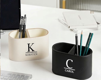 May include: Two personalized desk organizers. One is cream-colored with the initial "K" and the name "Kirsten". The other is black with the initial "C" and the name "Carol". Both have two compartments and hold pens and pencils.