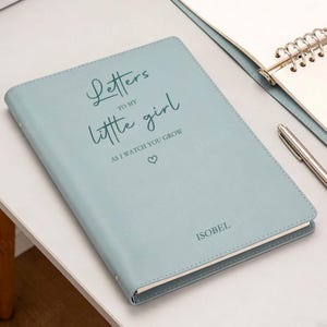 May include: A pale blue journal with the inscription "Letters to my little girl, as I watch you grow" and a heart symbol. The name "ISOBEL" is printed at the bottom. A pen and notebook are in the background.