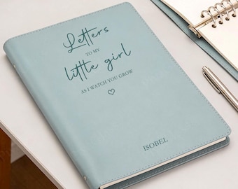 May include: A light blue journal with the text "Letters to my little girl, as I watch you grow" and a heart symbol. The name "ISOBEL" is printed at the bottom. A pen and notebook are in the background.