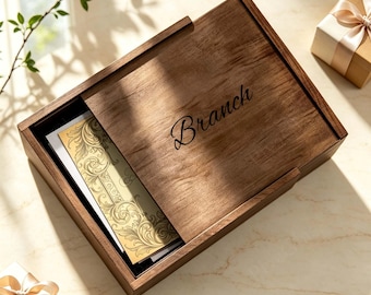 May include: A rectangular wooden box with a sliding lid featuring the word Branch engraved in black script. Inside the box, a cream-colored card with intricate gold filigree patterns is visible. Two small gift boxes with ribbons are nearby.