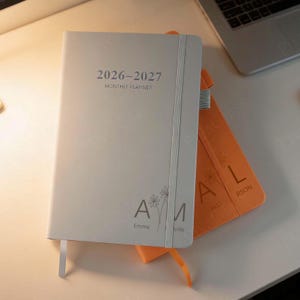 Op de afbeelding: Een lichtgrijze maandplanner 2026-2027 met de naam "Emma" en een bloemmotief. Een oranje planner met de naam "Alli" staat erachter. Beide hebben elastische banden en lint bladwijzers.