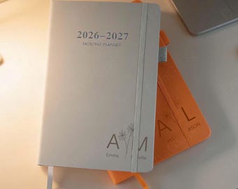 May include: A light gray 2026-2027 monthly planner with the name "Emma" and a floral design. An orange planner with the name "Alli" is behind it. Both have elastic bands and ribbon bookmarks.