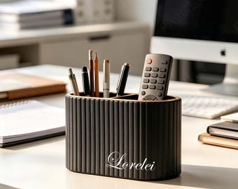 May include: A matte black oval desk organizer with a vertical ribbed texture and the name Lorelei printed in white script on the front. The organizer holds several pens, pencils, and a black remote control with multiple buttons.