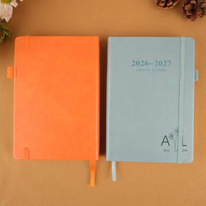 Op de afbeelding: Twee hardcover planners. E&eacute;n is oranje, de andere lichtblauw. De lichtblauwe planner heeft de tekst "2026-2027 MONTHLY PLANNER" en de initialen "A L" met een bloemmotief. Beide hebben lint-bladwijzers.