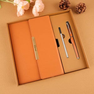 Könnte beinhalten: Ein orangefarbenes, ledergebundenes Tagebuch mit einem goldenen Metallakzent und dem eingravierten Wort "Liemi". Das Tagebuch befindet sich in einer braunen Schachtel mit zwei Stiften, einem silbernen und einem braunen.