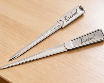 May include: Two silver letter openers with the name "Rachel" engraved on them. The letter openers are on a light brown wooden surface. The letter openers have a sleek, modern design.