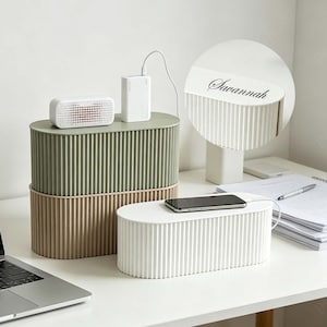 May include: Three cable tidy boxes in green, beige, and white, with a ribbed design. A phone rests on the white box, and a power bank and speaker sit atop the green box. The white box has the name "Savannah" printed on it.