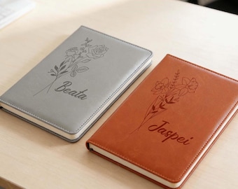 May include: Two personalized notebooks. One is gray with the name "Beata" and a floral design. The other is brown with the name "Jaspei" and a floral design. Both notebooks have a stitched border.