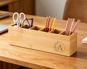 May include: A rectangular bamboo desk organizer with five compartments. The front features a circular engraved design with a leaf wreath, the letter A, and the name Amanda. It holds silver scissors, black glasses, and several colored pencils.