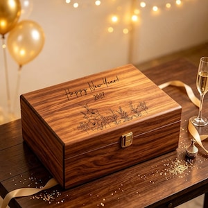 May include: A rectangular wooden box with a dark brown wood grain finish, featuring a gold-coloured metal latch. The lid is engraved with the text "Happy New Year! 2027" above a detailed line art illustration of various wildflowers and foliage.