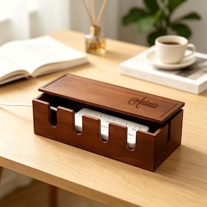 May include: A rectangular dark wood cable management box with a removable lid featuring the engraved name Antonia. The box has four U-shaped cutouts along the front edge for cords and contains a white power strip with multiple electrical outlets.