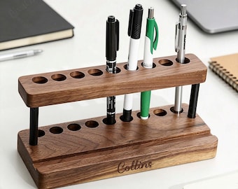 May include: A wooden pen holder with two tiers, holding several pens of various colors. The holder is made of dark wood and has the name "Collins" engraved on the front. The pens include black, white, and green pens. The holder is on a white surface.