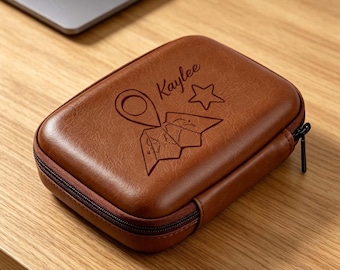 May include: A rectangular brown zippered case with a smooth, leather-like texture. The top surface features an engraved design including a map icon, a star, a location pin, and the name Kaylee written in a cursive script font.