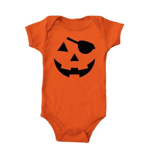 May include: An orange baby bodysuit featuring a black jack-o'-lantern face. The design includes a triangle nose, a curved mouth, and an eye patch. The short-sleeved bodysuit has snap closures at the bottom.