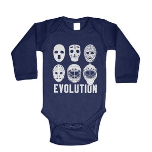 May include: A navy blue long-sleeved baby onesie with a white graphic of hockey goalie masks, showing the evolution of the mask design. The text "EVOLUTION" is printed below the masks.
